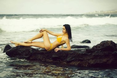 Sarı mayolu güzel kız sahilde kayalıklarda oturuyor.