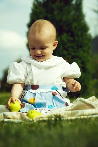 スタイリッシュで幸せな笑みを浮かべて面白い小さな赤ちゃん女の子の自然な草の背景 画像の垂直方向に緑のリンゴといっしょにピクニックに座っているが屋外ドレスします — ストック写真