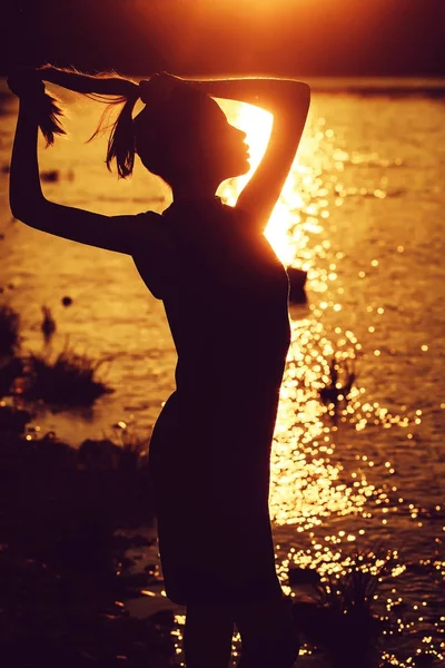 pretty sexy female silhouette of woman or girl with raised hands outdoor over dramatic dark sky with clouds and sea or ocean water on evening natural sunset background