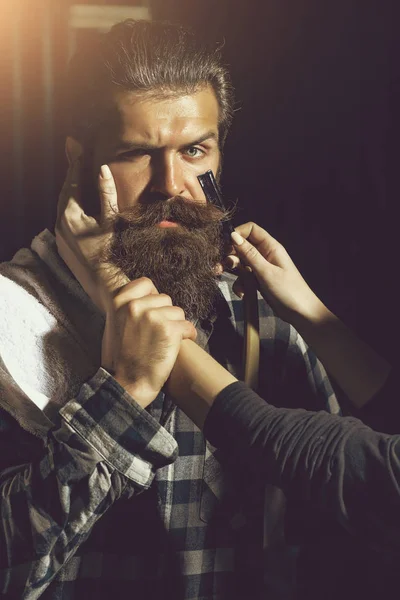 Female hands shave bearded man, handsome hipster, brunette with beard and moustache. Woman works with vintage razor on dark background