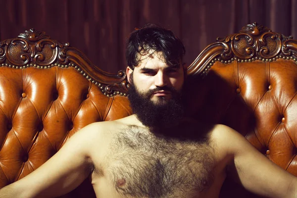 One handsome serious cool young adult man with long black lush beautiful beard moustache and bare hairy chest sitting on leather couch looking forward indoor on studio background, horizontal picture