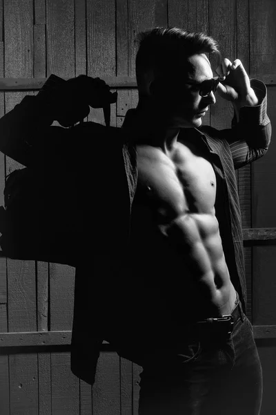 セクシーな深刻な官能的な筋肉マッチョな若者裸の胴体とスタイリッシュな髪シャツ サングラスと屋内木製の背景画像の垂直方向に立っているバッグ — ストック写真