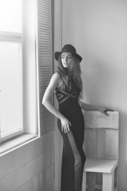 Kız ya da güzel moda kadın siyah giyen uzun sarı saçları ile şapka hoş ve ayakta ahşap sandalye ve pencere beyaz duvar arka plan üzerinde elbise