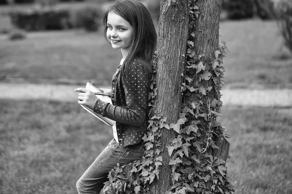 Chica con cuaderno y pluma cerca del árbol — Foto de Stock