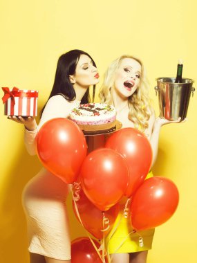 İki sevimli seksi genç mutlu sarışın ve esmer kadın doğum günü pastası mumlar yığın konfeti ve şarap şişe kırmızı balonlarla demet yakınındaki ile Studio Sarı zemin, dikey fotoğraf üzerinde tutan