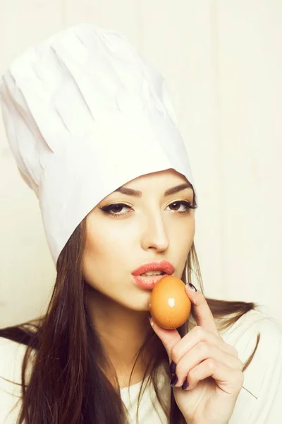 One beautiful sensual young female cook in professional white uniform of hat and apron cooking homemade food holding egg near face in kitchen on wooden background, vertical picture