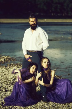 Yakışıklı adam uzun saçlı mor elbiseler yakınındaki su güneşli gün açık erkek bacaklar kucaklayan iki genç güzel kadın ile ayakta beyaz gömlekli sakallı
