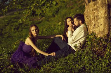 Yeşil çim güneşli açık ağaç yakınındaki mor elbise iki genç güzel kadınlarla oturup sigara Beyaz gömlekli sakallı yakışıklı adam