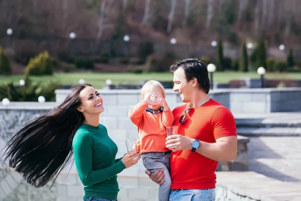 Happy family couple of young pretty woman and handsome man in park with son holding glasses of water and smiling