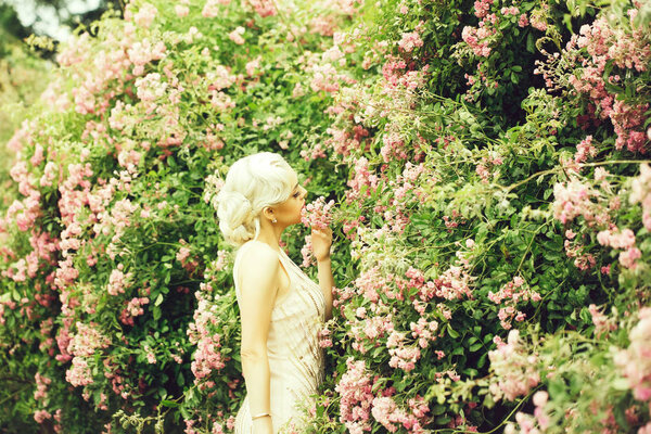 Young girl with blonde hair fashion makeup on pretty face in beautiful cream-colored dress posing near pink rose bush in bloom