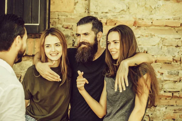 Couple of handsome bearded man with long beard and stylish hair and blonde pretty cute sexy women or girls embracing and smiling with friend on summer sunny day on stony wall background