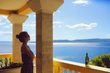 Sevimli kadın şarap yakınındaki deniz manzarası ile cam korkuluk açık terasta üzerinde bakarak.