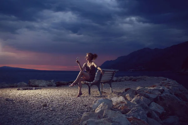很性感的女人或女孩与酒杯坐在长凳上户外在黑沉沉暮色里上空云在海滩上与大海或海洋水晚上自然背景上 — 图库照片