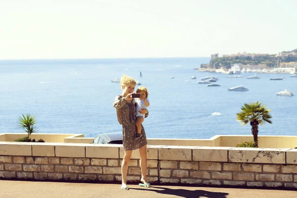 Monte Carlo, Monaco - September 20, 2015: mother woman young beautiful with son takes selfie on smart phone promenade on summer sunny day at blue sea side on seascape background, horizontal picture