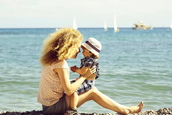 ペブル ビーチ青い海海岸を楽しんで美しい彼女はひざまずいて日当たりの良い夏の日一緒に座っての息子と母親の若い女性がぼやけて海背景 画像の水平方向 — ストック写真