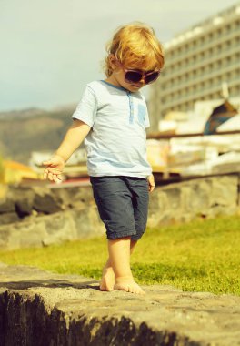 Sarı saçlı mavi tshirt ve şort ile güneş gözlüklü şirin moda bebek çocuk yalınayak güneşli yaz günü doğal zemin üzerine taş duvara yürür