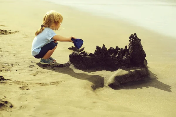 かわいい男の子は青い シャツとショート パンツでブロンドの髪のポニーテール シースケープ背景の日当たりの良い夏の日の海ビーチにシャベルで砂の城を構築します — ストック写真