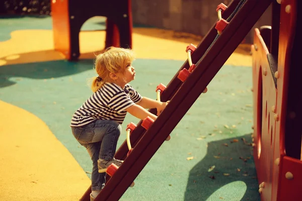 シャツとジーンズで金髪のかわいい男の子が遊び場の背景の日当たりの良い夏の日の赤い木製スライド ボードのはしごを登る — ストック写真