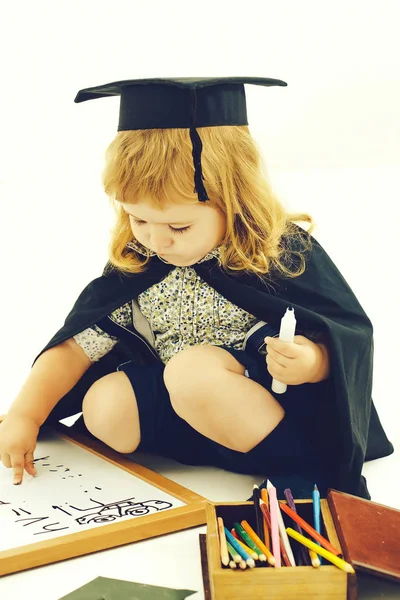 Little boy child in black academic gown and squared hat playing with drawing school board holding marker near box with colored pencils isolated on white background