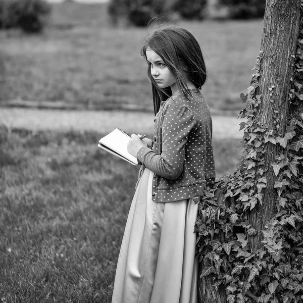 Девушка в платье с блокнотом и ручкой рядом с деревом — стоковое фото