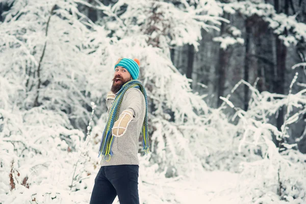 Skincare, beard care in winter. Hipster in thermal jacket, hat, scarf, beard warm in winter. Snow fight, sport, rest. Bearded man smile with snowballs in snowy forest. Temperature, freezing, cold snap