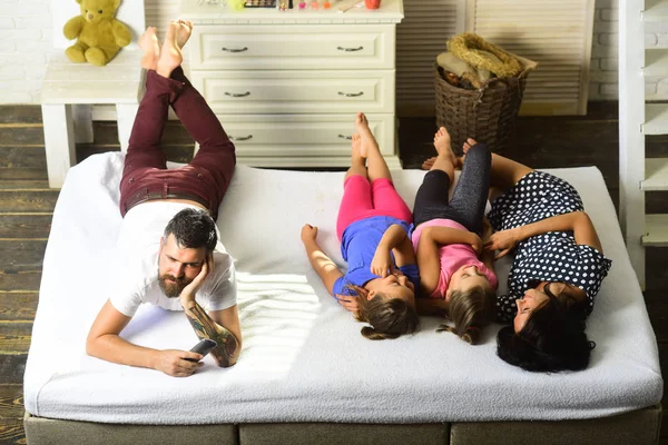 Loving family and entertainment concept. Family with cheerful and happy faces at home. Mother, father and daughters with tv remote lying on bed. Man, woman and adorable kids watching tv in apartment.