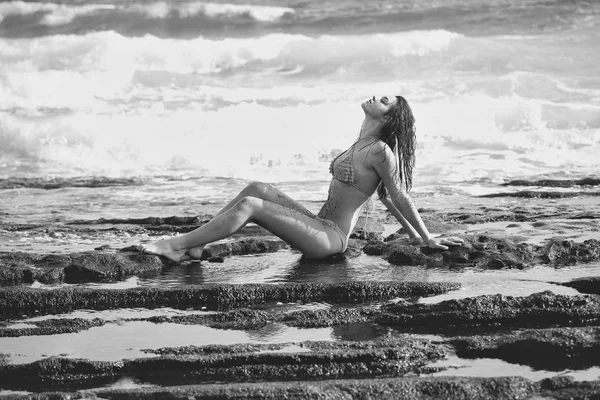 Красива дівчина в сексуальному жовтому купальнику, що сидить на кам'янистому пляжі — стокове фото