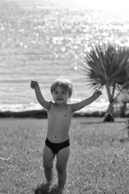 Kırmızı şort sarışın saçlı şirin neşeli bebek çocuk elleri açık havada mavi deniz zemin üzerine yeşil çimenlerin üzerinde pastoral, güneşli, yaz gününde yükseltir