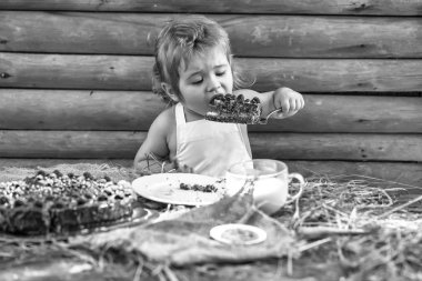 Beyaz önlük içinde şirin küçük çocuk ahşap arka plan açık havada masaya, fincan süt ve meyve kek yiyor