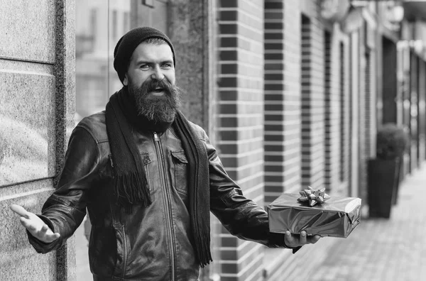 bearded man, long beard, brutal caucasian hipster with moustache holding blue present or gift box, has happy smiling face, in black leather jacket, hat, scarf, unshaven guy at building outdoor