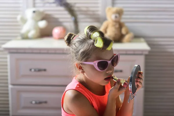 Little girl paints lips with serious face sitting on bed in childroom. Child wears sunglasses and takes mothers cosmetics. Girl with curlers and lipstick in room. Makeup and kids fashion concept.