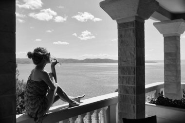 yaz günü doğal zemin üzerine deniz ya da okyanus su üzerinde seyir cam beyaz şarap içme sütunlarla balkonda oturan güzel kadın