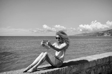 Genç güzel sarışın uzun kıvırcık saçlı kerbstone üzerinde oturan ve deniz manzarası arka plan, yatay resim üzerinde mavi deniz tarafında güneşli gün yaz akıllı telefon selfie alarak 