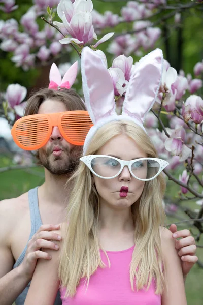 couple in love making fish lips, face grimace in glasses and rosy, bunny ears. Pretty girl or cute woman and handsome man or macho on blossoming, floral environment. Spring. Easter, celebration