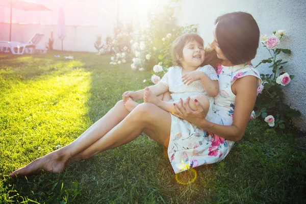 Nurturing and future. Mothers day concept. Woman with baby girl sitting on green grass. Family love and care. Mom and child smiling at blossoming rose flowers.