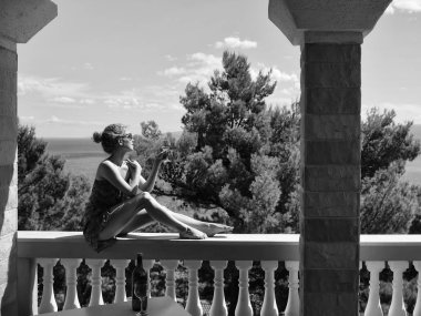 yaz günü yeşil çam doğal zemin üzerine deniz ya da okyanus su üzerinde seyir cam beyaz şarap içme sütunlarla balkonda oturan güzel kadın