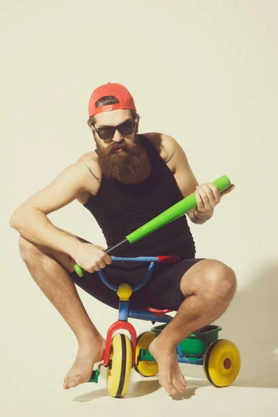 Bearded angry man holds green baseball bat on bicycle toy — Stock Photo, Image