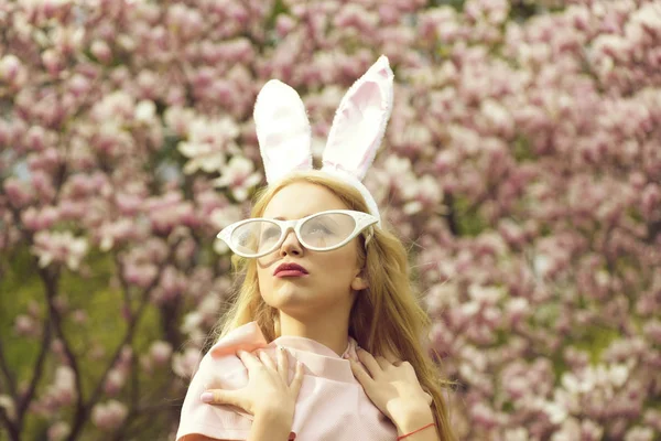 漂亮女孩或可爱的女人 兔子耳朵与滑稽的眼镜 长长的金发头发摆在模糊 花艺环境公园里开花树木的粉色上衣时尚模型 复活节 — 图库照片