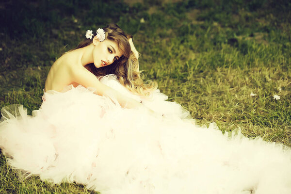 Beautiful young woman in pink glamour dress with long curly hair sitting on green grass in spring sunny day