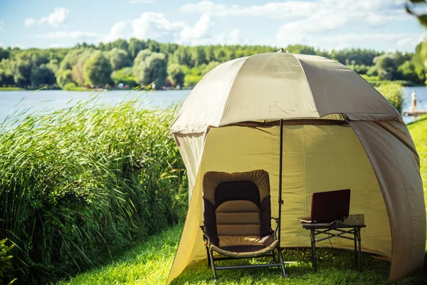 Fishing tent with armchair, laptop on summer landscape