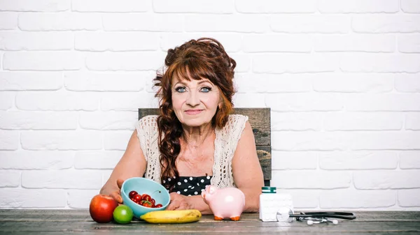 Senior vrouw met spaarvarken, fruit, geneeskunde, stethoscoop aan tafel — Stockfoto