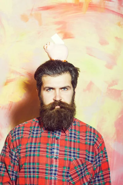 Homem de barba franzido segurando rosa banco de cerâmica porquinho na cabeça — Fotografia de Stock