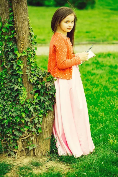 Menina em vestido com caderno e caneta perto da árvore — Fotografia de Stock
