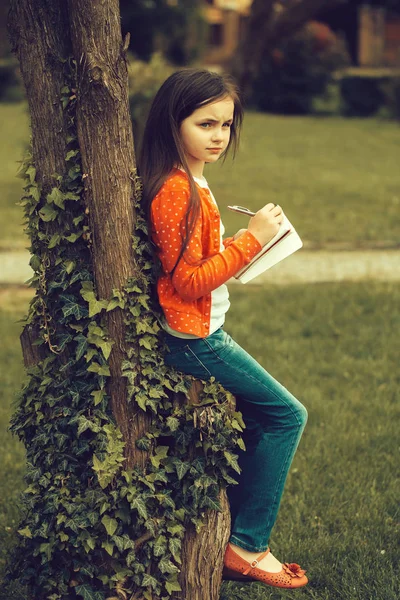 ノートブックとツリーの近くでペンを持つ少女 — ストック写真