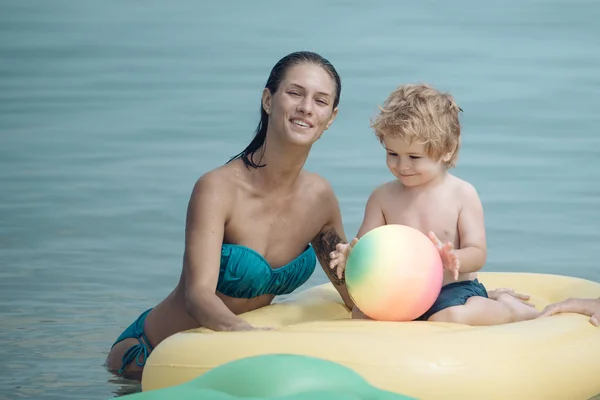 可爱的儿童男孩与坐在空气床垫菠萝形状, 在海洋, 海。母性观念。母亲站在附近, 用儿子推床垫。带着笑脸的妈妈和孩子在海里共度时光. — 图库照片