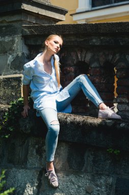 Kadın gömlek ve kot taş duvarda, moda otur. Uzun sarı saçlar, saç modeli üzerinde güneşli açık olan kadın. Manken makyaj yüz, güzellik. Kız Stil, güzellik, göz ve vogue.