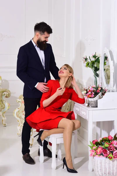 Elegant couple near toilet table, lady busy with make up, luxury interior on background. Man in suit, woman holds lipstick. Couple in love preparing to go out, elite event. Elegant outfit concept.