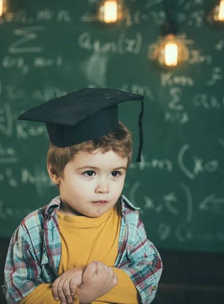 Bambino intelligente in berretto laureato su faccia seria, timido, tiene le mani incrociate. Bambino, in età prescolare o il primo ex, lavagna su sfondo, sfocato. Concetto di laurea. Ragazzo sembra carino in cappello accademico quadrato . — Foto Stock