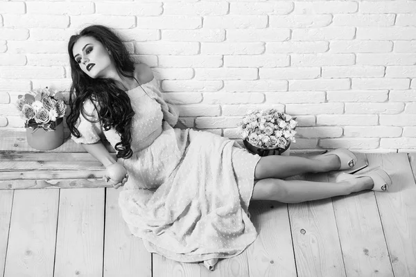 pretty sexy woman or girl with cute face and long brunette hair has fashionable makeup with red lipstick in dress near pink rose flowers bouquet in box holds green apple on wood floor and brick wall