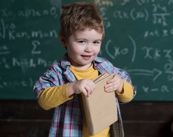 Smart child on smiling face, holds book in hands. Kid, preschooler or first former, chalkboard on background, defocused. Studying concept. Boy looks cute with book, kid likes to study.